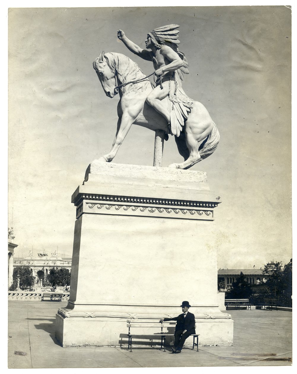 Lodret, sepia -fotografi, der viser en statue af en amerikansk indianer siddende på en hest oven på en stor piedestal. Der sidder en mand i mørkt jakkesæt og hat på en bænk ved sokkelbunden af Unknown