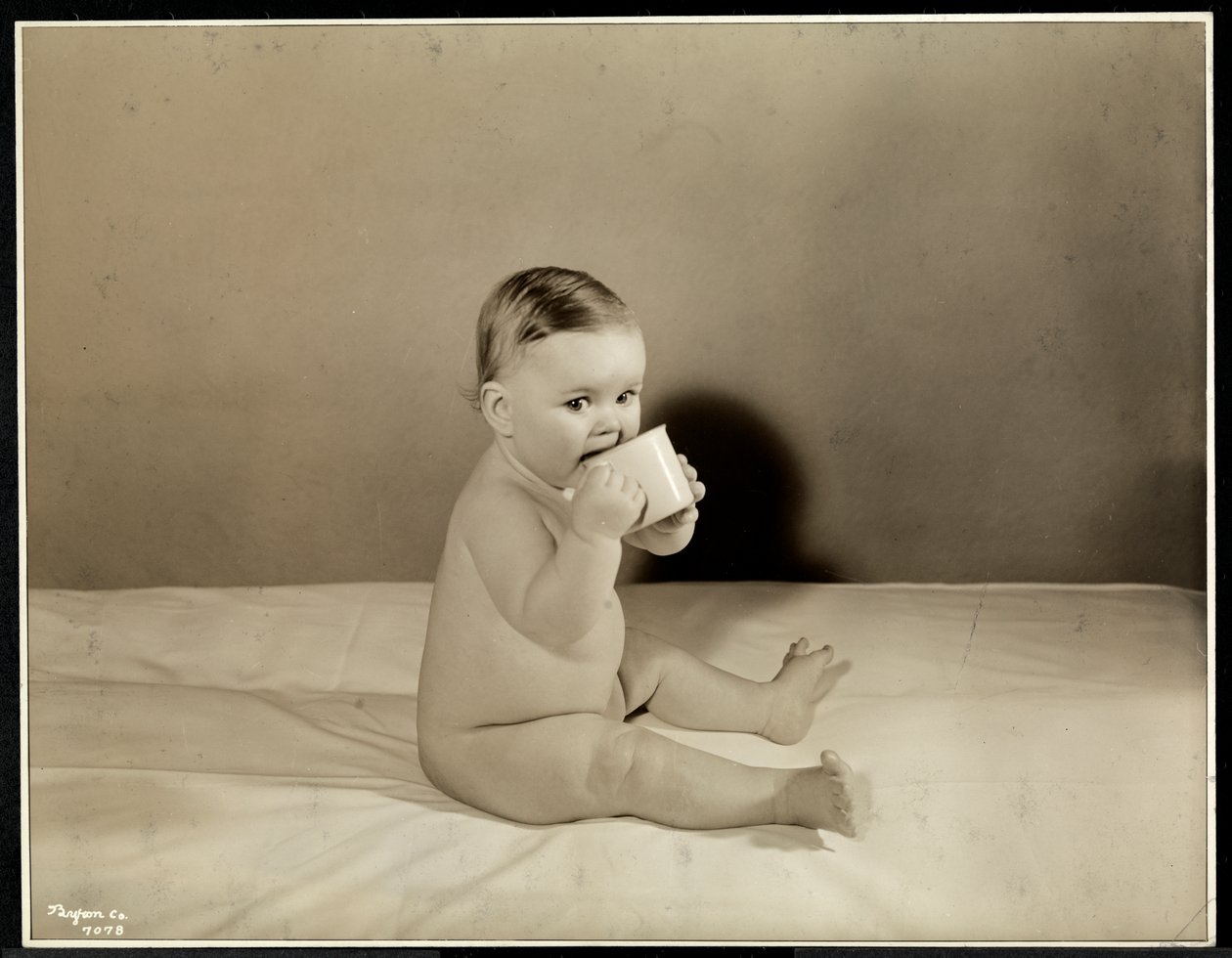 En baby, der sidder oppe i sengen og drikker af en kop hos Maternity Center Association på 70th Street East River, New York, 1935 (sølv gelatinetryk) af Byron Company