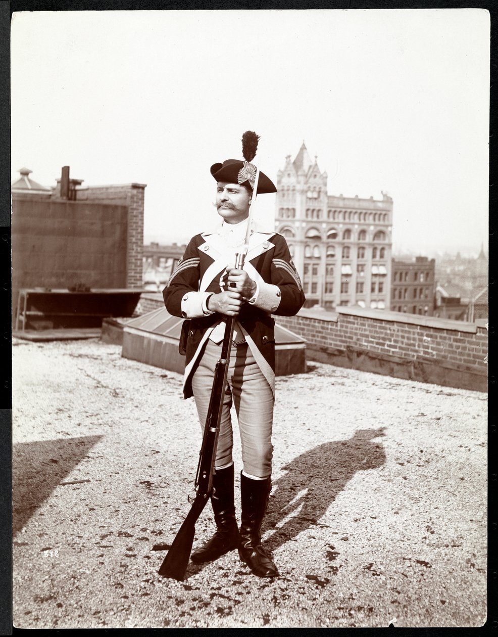 En mand på et hustag klædt i traditionel militæruniform med en riffel i hånden, 1895 (sølvgelatineprint) af Byron Company