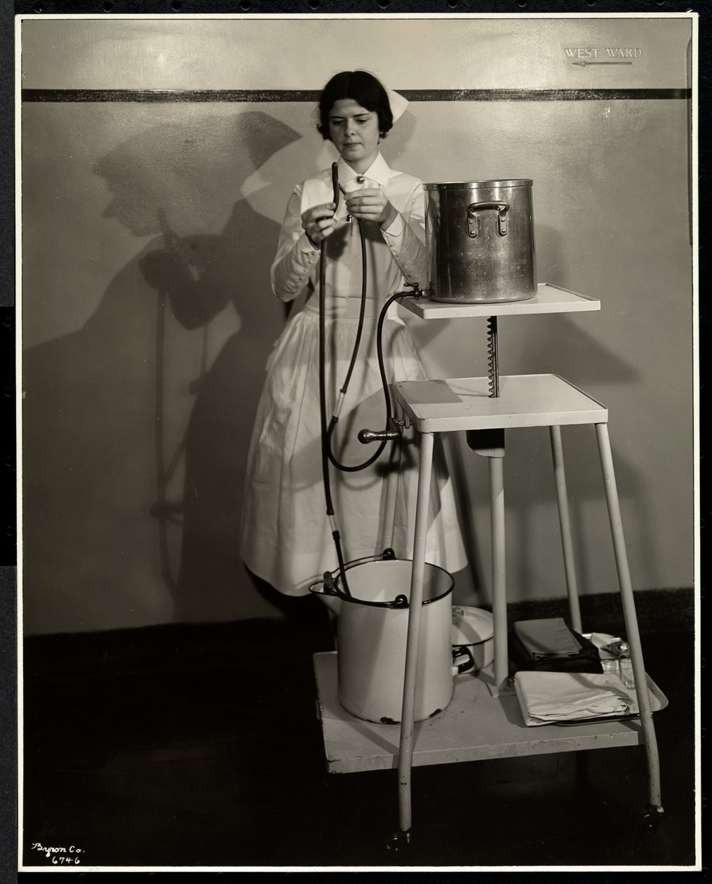 En sygeplejerske demonstrerer et specielt apparat med rør og forsynings- og affaldsspande på Sloan Maternity Hospital, Broadway og 168th Street, New York, 1932 (sølv gelatinetryk) af Byron Company