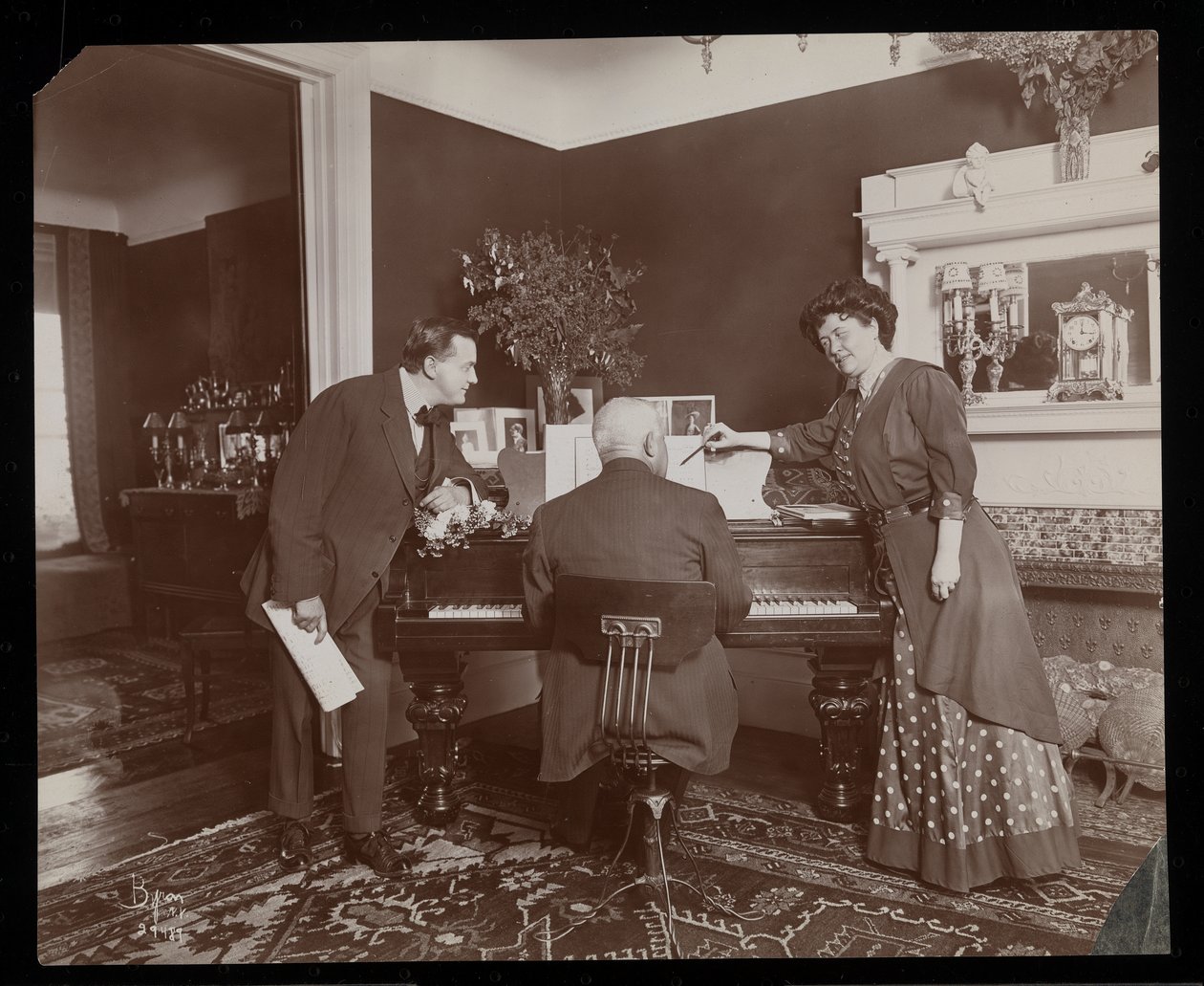 Clara De Regand peger på et partitur, mens en mand spiller klaver, 1910 (sølv gelatinetryk) af Byron Company