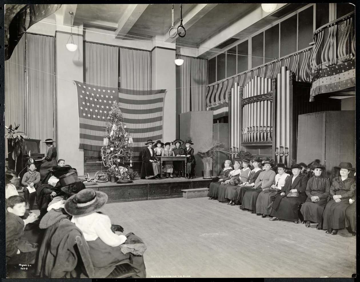 Folk i et auditorium i New York Association for the Blind med juletræ og folk, der muligvis opfører et sketch på scenen med orgel, 1920 (sølv gelatinetryk) af Byron Company