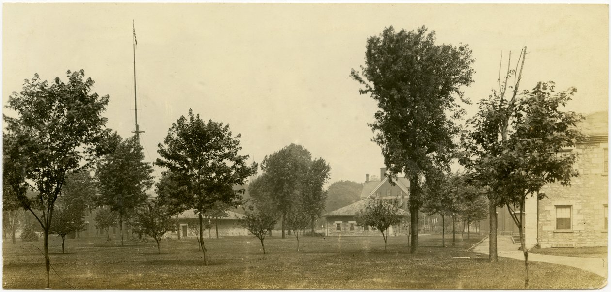 Vandret, sepia -fotografi, der viser en gruppe murstensbygninger, der grænser op til et åbent grønt område med træer spredt overalt. Nogle af murstensbygningerne ser ud til at være huse af Carl Michel