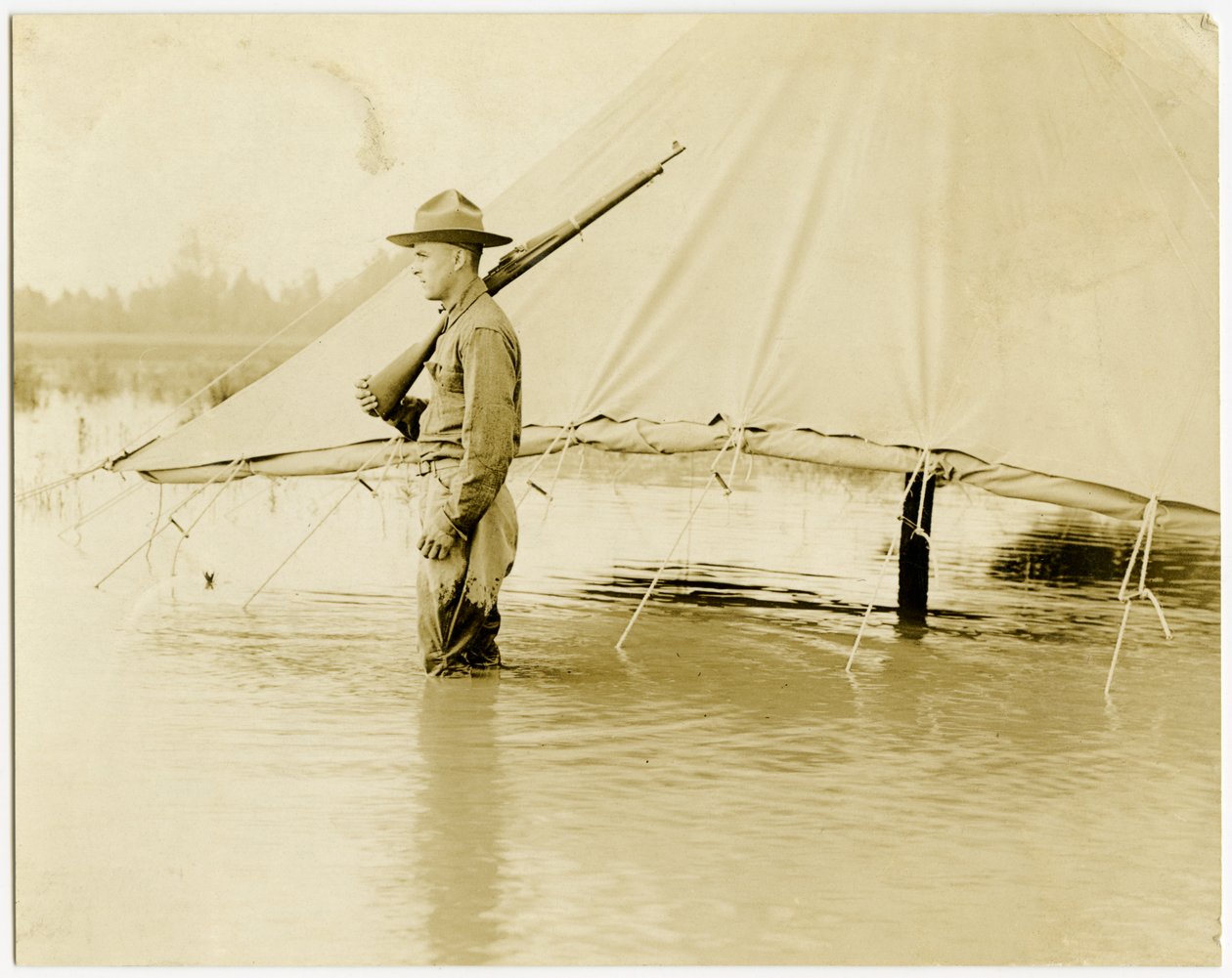 Vandret, sepia -fotografi, der viser en uniformeret soldat, der står vagt i et oversvømmet felt. Soldaten står ved siden af et telt, som har siderne rullet op og sikret over vandlinjen af Carl Michel