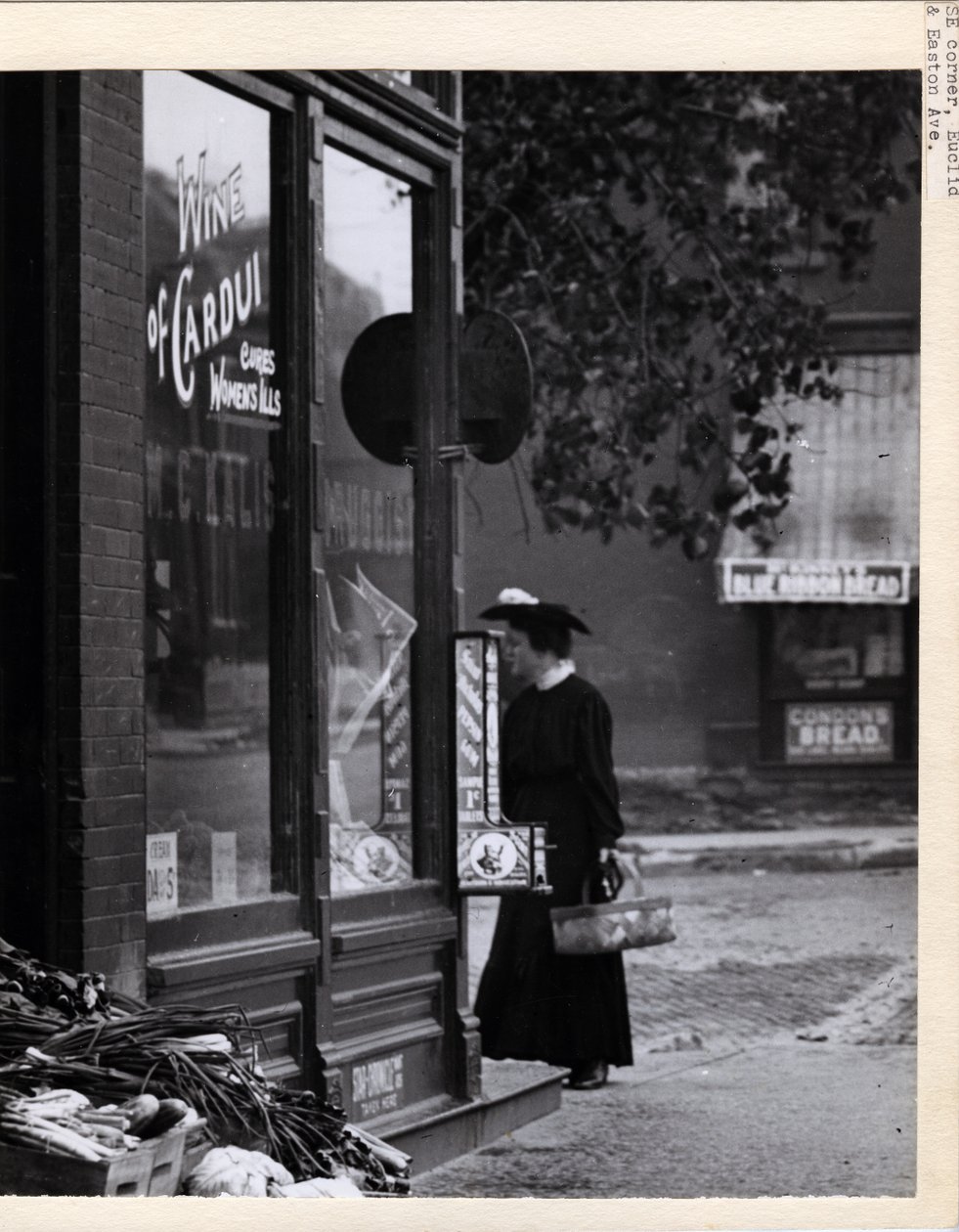 Lodret, sort og hvidt fotografi, der viser en kvinde, der holder en kurv, mens hun stod foran W. C. Kalis Drugstore -indgangen på 4878 Easton Avenue af Charles Clement Holt