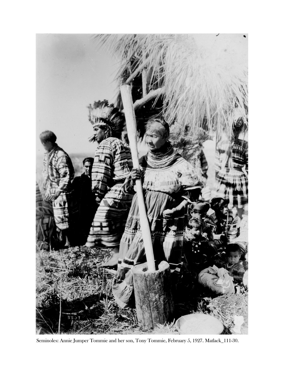 Annie Jumper Tommie brugte en morter og støder til at lave majsmel, og hendes søn, Tony Tommie, iført en ørnefjerhjelm, 5. februar, 1927 af Claude C. Matlack