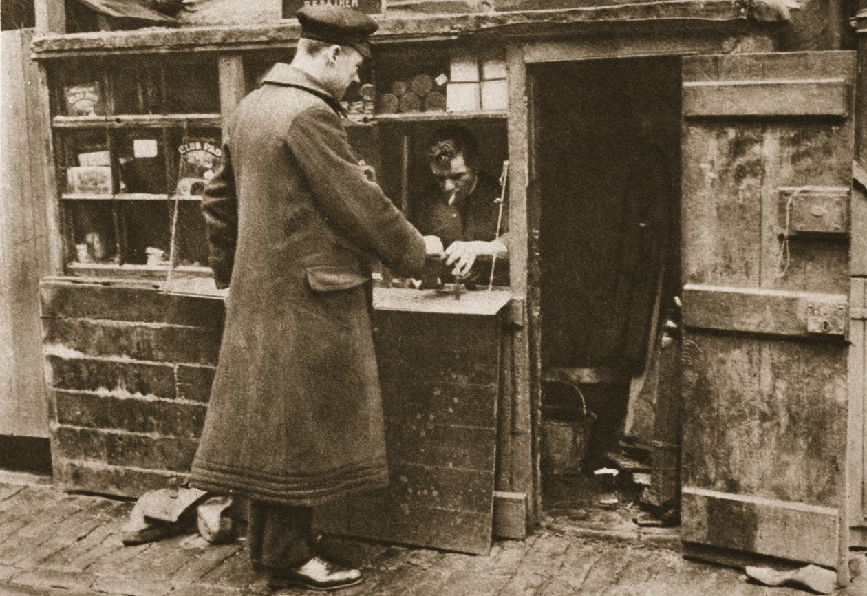 En skomager, etableret i en provisorisk kabine i en brostensbelagt Mayfair mew, tager støvler ind fra en chauffør fra Wonderful London, udgivet 1926-27 (fotogravure) af English Photographer