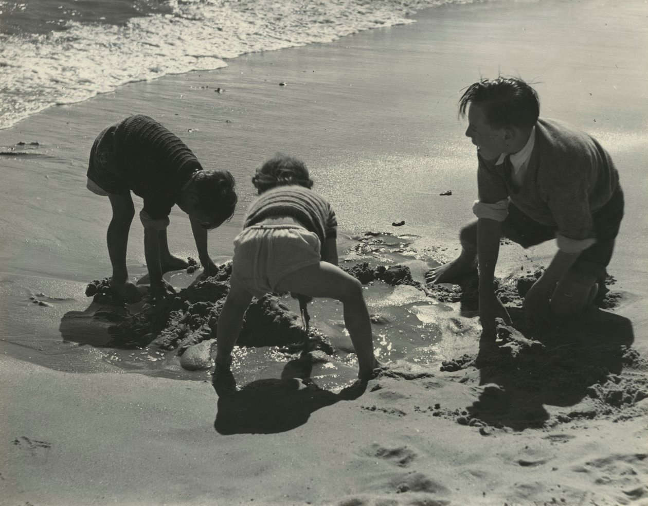 Tre børn leger med sand på stranden. af Godfrey Macdominic