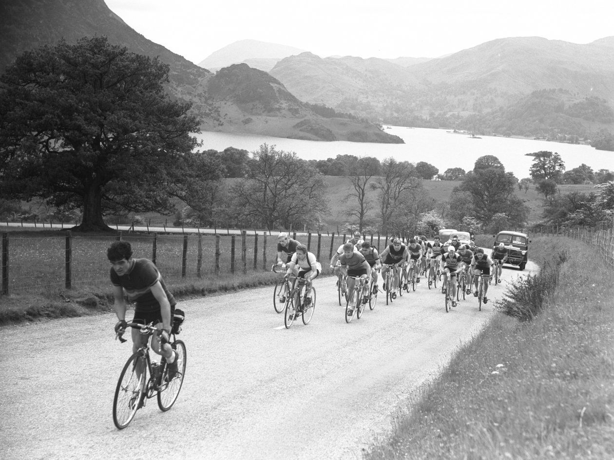 Gruppe af cyklister på vej op ad en vej efterfulgt af biler, sø og fjelde i baggrunden af Joseph Hardman