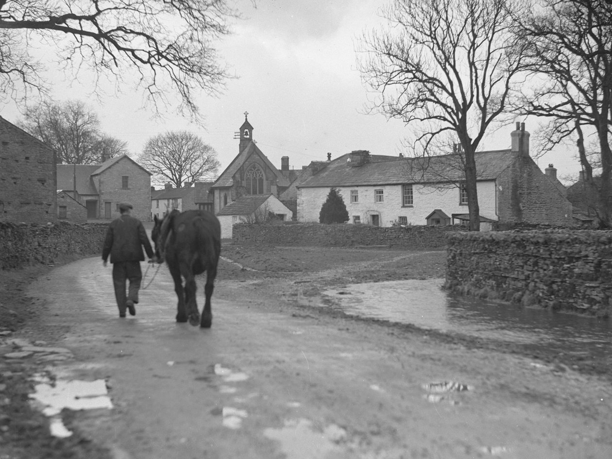 Mand fører hest ned ad vej væk fra kameraet mod landsbybygninger og kirke; arkitektur; religiøs; kirkelig; af Joseph Hardman