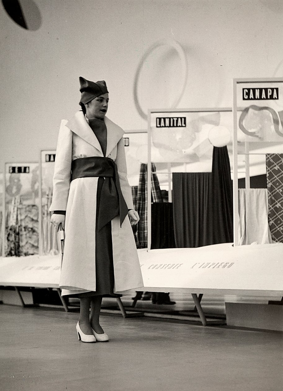 Model i en overfrakke skabt af couture-huset "Lamma". Artiklen er samlet i taljen af et bredt skær og afsluttet med en blød hat. Fotografiet er taget i en hal, der bruges til udstilling af stoffer. (s/h foto) af Luis Bogino