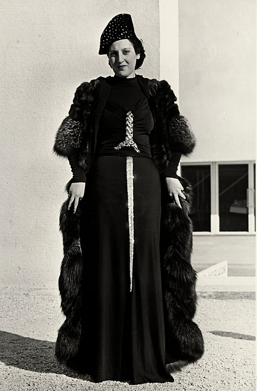 Model i aftenkjole skabt af det italienske couture-hus "Dragoni". Artiklens skrifttype har en prangende pailletdekoration i form af et sværd. En hat og pelsoverfrakke fuldender ensemblet. (s/h foto) af Luis Bogino