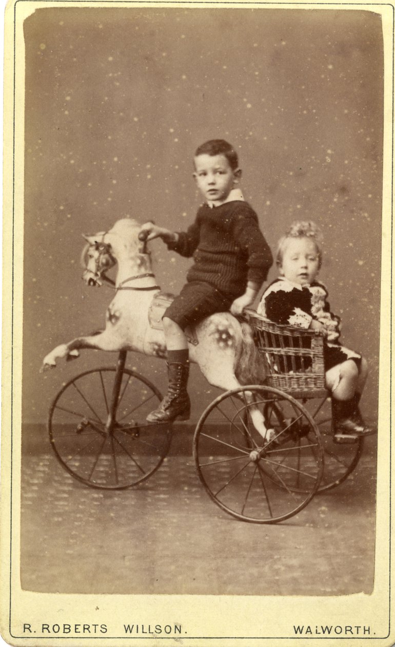 Ung dreng på gyngehest med ung pige i fletvogn bagved, 1880