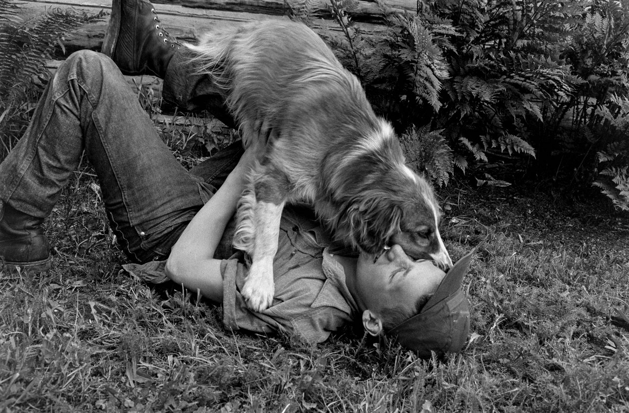 En ung mand leger med sin hund uden for deres bjælkehytte i Palmer, Alaska. af Ted Spiegel