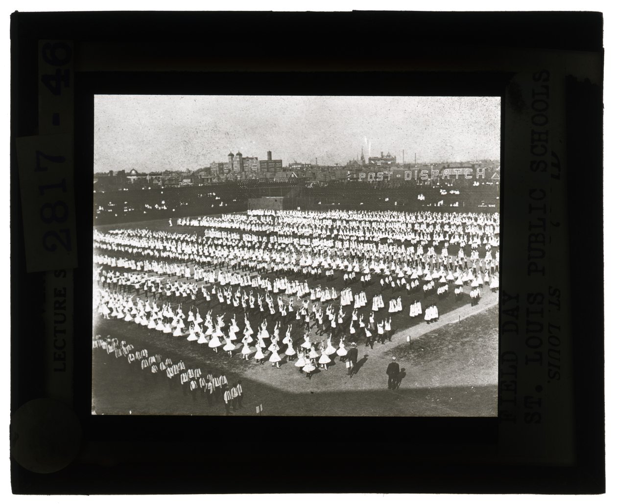 Vandret, sort -hvidt foto af hundredvis af skolebørn stillede op i formation til Field Day -parade og fest. Alle pigerne i hvide kjoler og drengene i hvide skjorter og slips med mørke bukser af Unbekannt Unbekannt