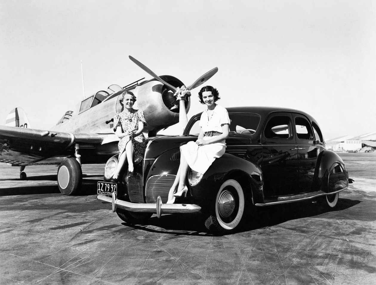 Californien: 1939 To kvinder sidder på skærmene af en Lincoln Zephyr sedan 1939 i en lufthavn med et enkeltmotors fly parkeret bag sig (s/h foto) af Unbekannt