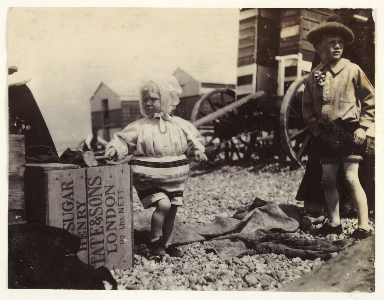 To børn ved bademaskiner, c 1895, Et øjebliksbillede af to børn ved havet, stående blandt en gruppe bademaskiner på stranden af Unbekannt