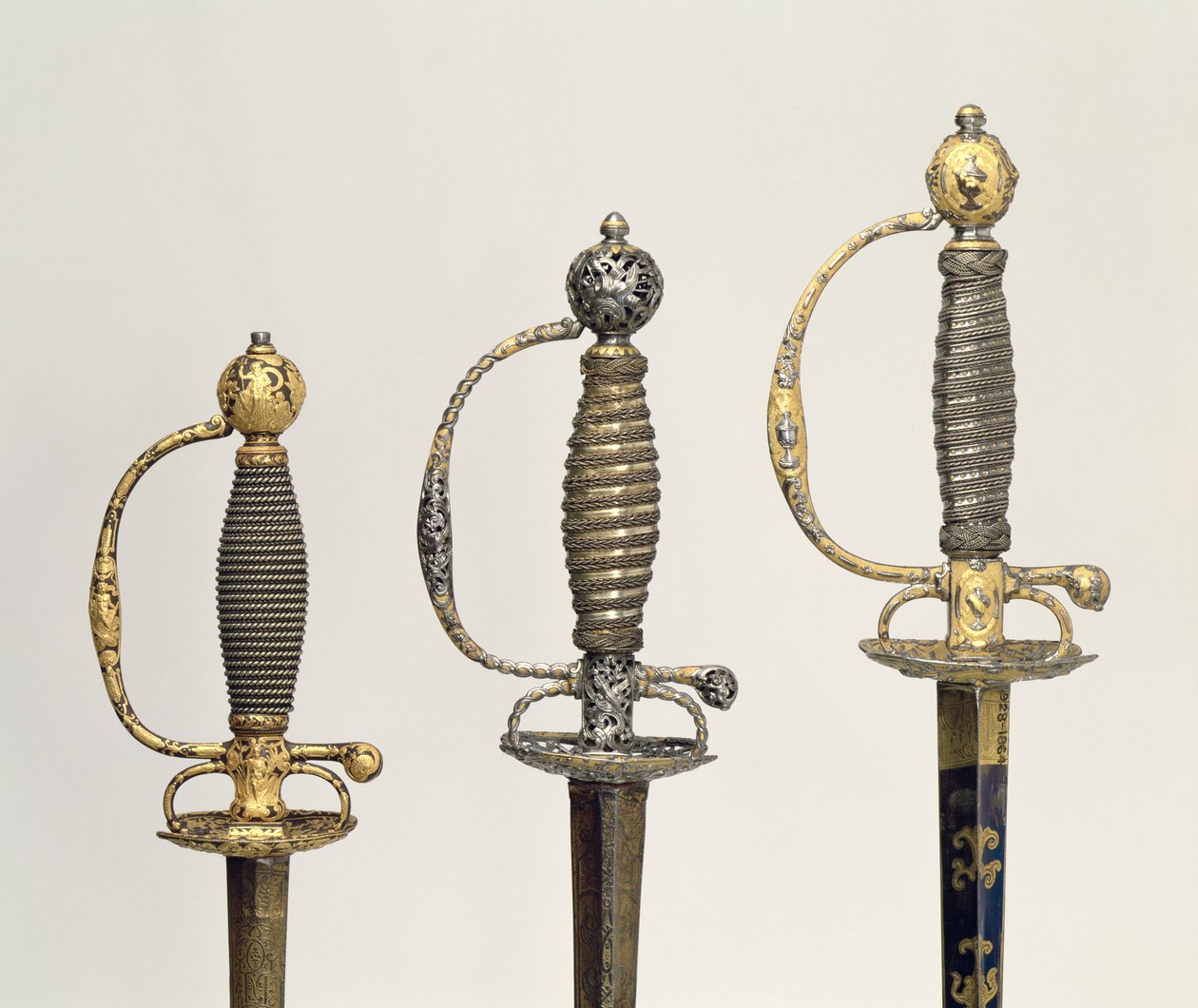 Hilt af tre franske sværd: et af stål, blåt og overtrukket med mønstre i jaget guld, ca. 1730; en af stål gennemboret og mejslet og slebet af matteret guld, c.1760; en af mejslet stål, gr af Unbekannt Unbekannt