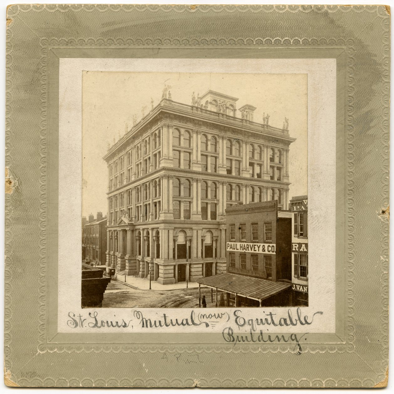 Fotografi af en seks etagers bygning med figurer langs toppen. Der er andre bygninger synlige til hver side. Bygning kaldes også St. Louis Life Insurance Building af Unknown