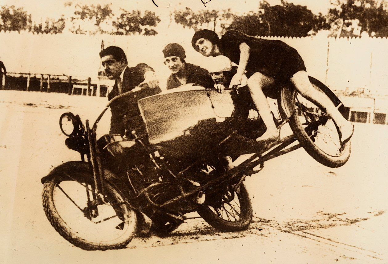 En mand viser sig frem på en motorcykel med en sidevogn, der bærer tre unge kvinder. Sidevognen løfter sig fra jorden, da de drejer. af Unknown photographer