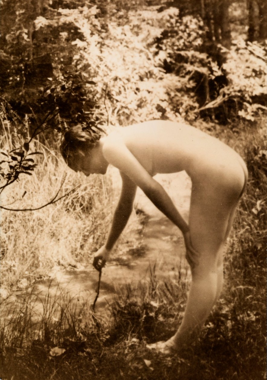 En ung kvinde, nøgen i skoven, ses i profil, mens hun bøjer sig for at lægge en gren i en bæk. af Unknown photographer