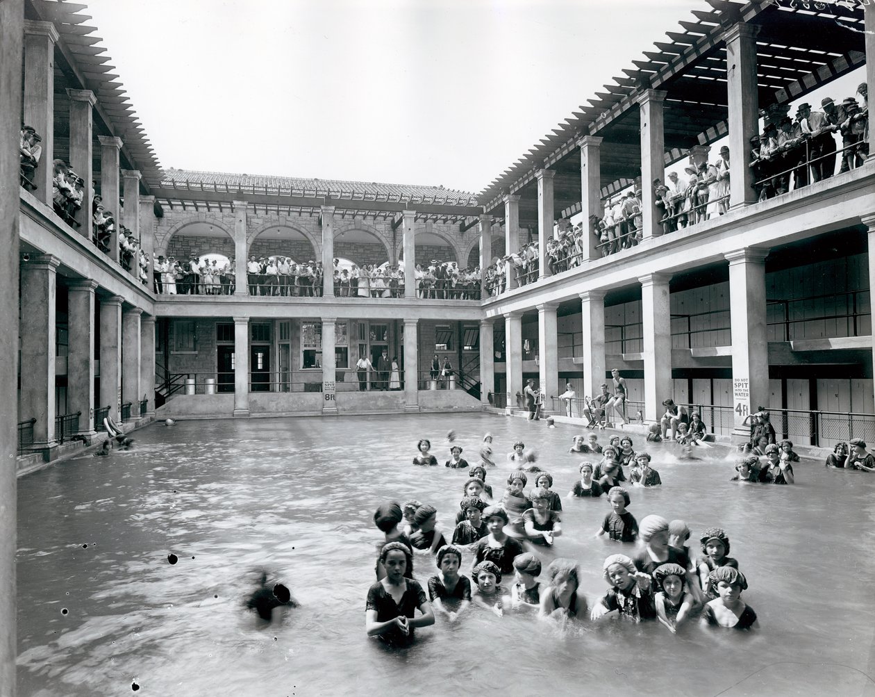 Vandret, sort / hvidt fotografi af en swimmingpool med to etagers balkon understøttet af søjler. Tilskuere ser svømmerne fra balkonen på andet niveau af William G. Swekosky