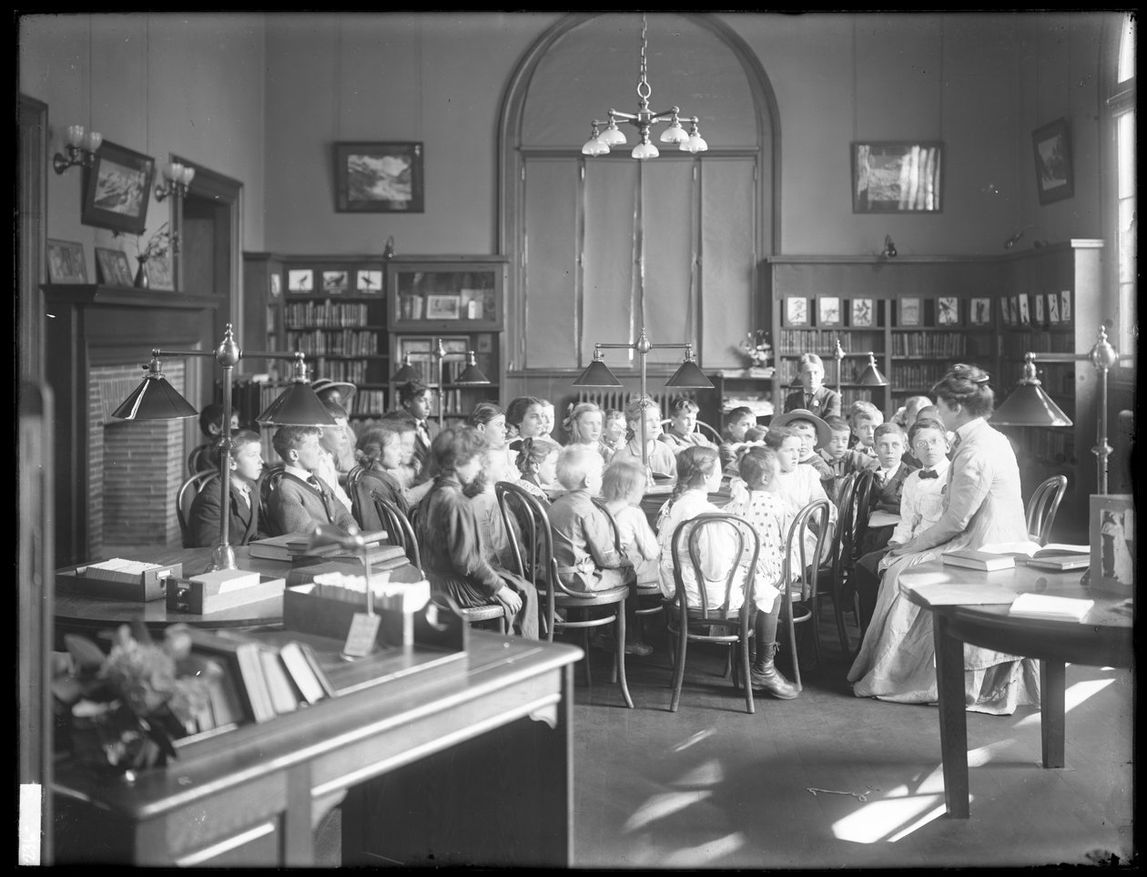 Børn lytter til en kvinde i læsesalen i en uidentificeret afdeling af Queens Borough Public Library, ca. 1910 af William Davis Hassler