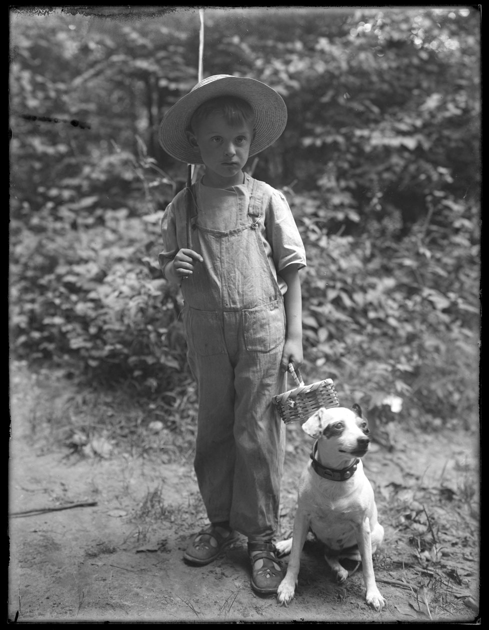 William Grey Hassler i stråhat og overalls, med fiskestang og kurv, poserede i skoven med en lille hund (Bessie), 20. august 1911 af William Davis Hassler