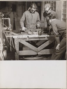 [Tre mænd med beskyttelsesbriller svejser et stykke metal på et bord].