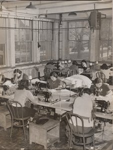 [Kvinder sidder ved skriveborde og arbejder på symaskiner].