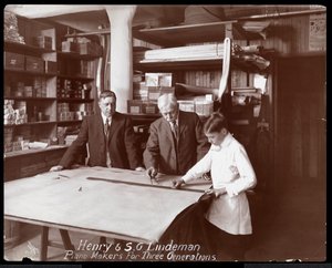 Portræt af Henry og S.G. Lindeman med deres søn eller barnebarn stående over butikstegninger til et klaver, 1910 (sølv gelatinetryk)