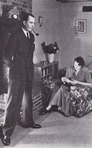 Hustru læser, mens mand varmer ryggen mod ilden, ca. 1950 af English Photographer