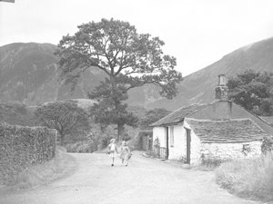 To unge piger, der holder hinanden i hånden med kviste af rævehaner, går ned ad vejen forbi et sommerhus mod kameraet. af Joseph Hardman