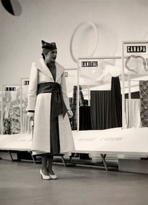 Model i en overfrakke skabt af couture-huset "Lamma". Artiklen er samlet i taljen af et bredt skær og afsluttet med en blød hat. Fotografiet er taget i en hal, der bruges til udstilling af stoffer. (s/h foto) af Luis Bogino