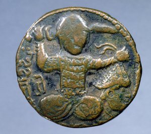 Forside af mønt (bronze) forestillende tyrker med hjelm, der sidder overskrævs med et sværd bag hovedet i højre hånd og et hjelmklædt afskåret hoved i venstre hånd. præget af Husam al-Din Yuluq Arslan (1184-1201)
