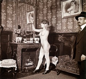 Gådefuldt fotografi, der viser en ung kvinde helt nøgen (bortset fra sko og strømper), som friserer sit hår foran et spejl i nærvær af en mand, der klæder sig på og varmer med en hat. Man kunne tro, at det var et foto taget på et bordel, men dette hyp af Unbekannter Künstler