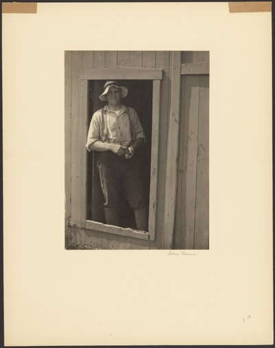 [Mand med stråhat står i døren til en lade og holder en tobakspose]. af Doris Ulmann