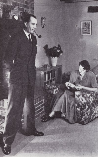 Hustru læser, mens mand varmer ryggen mod ilden, ca. 1950 af English Photographer