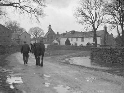 Mand fører hest ned ad vej væk fra kameraet mod landsbybygninger og kirke; arkitektur; religiøs; kirkelig; af Joseph Hardman