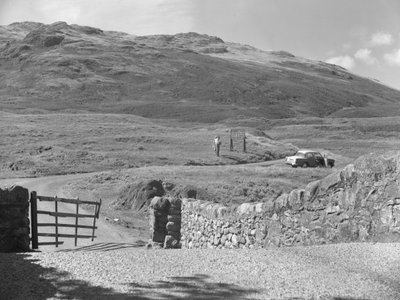 Udsigt til vejen, der går over Hard Knott Pass, to mennesker, der står ved et vejskilt, og en parkeret bil i græsrabatten af Joseph Hardman