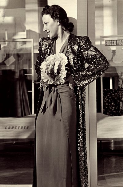 Model i aftenkjole skabt af det italienske couture-hus "Dragoni". Artiklen er pyntet med en imponerende blomst og suppleret med en overfrakke med pailletter. (s/h foto) af Luis Bogino