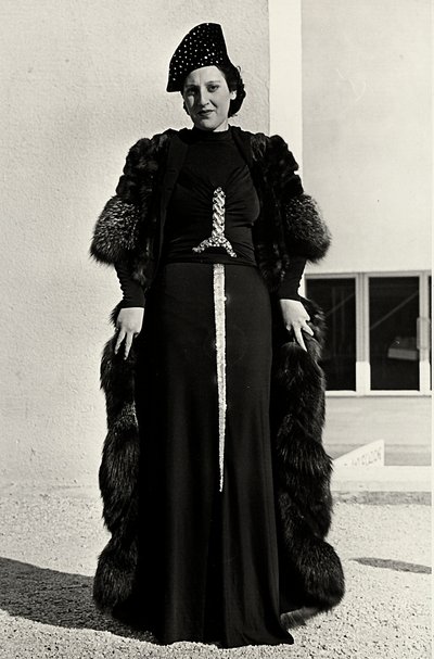 Model i aftenkjole skabt af det italienske couture-hus "Dragoni". Artiklens skrifttype har en prangende pailletdekoration i form af et sværd. En hat og pelsoverfrakke fuldender ensemblet. (s/h foto) af Luis Bogino