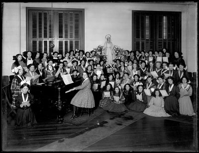 Gruppeportræt af en pigekor-klasse samlet omkring et klaver med noder, sandsynligvis på det romersk-katolske orphan asylum i Kingsbridge, Bronx, ca. 1911 af William Davis Hassler