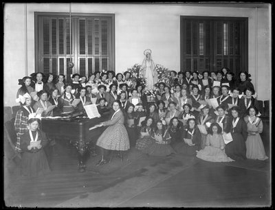 Gruppeportræt af en klasse for pigekor samlet omkring et klaver med noder, sandsynligvis på det romersk-katolske orphan asylum i Kingsbridge, Bronx, ca. 1911 af William Davis Hassler