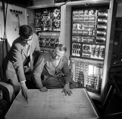 Ingeniører diskuterer planer foran det elektroniske kontrolpanel, de har bygget af Frederick (attr. to) Barnard