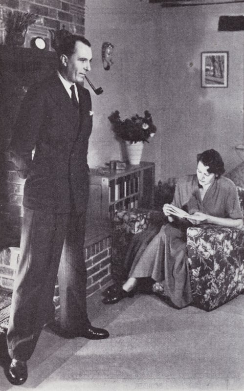 Hustru læser, mens mand varmer ryggen mod ilden, ca. 1950 af English Photographer
