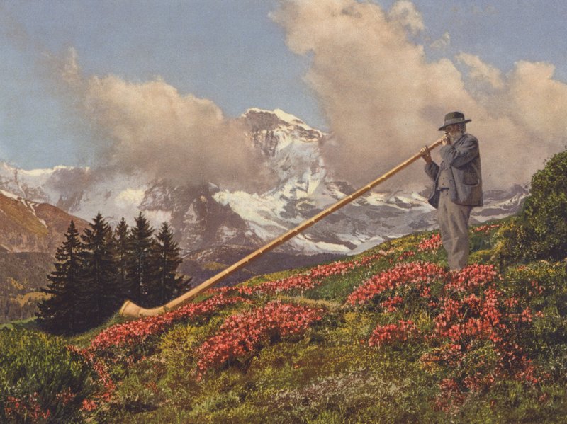 Schweiz: En alpehornsblæser, Jungfrau i baggrunden (farvefoto) af Jean Gaberell