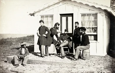 Fem borgerkrigssoldater samlet på snavset veranda uden for hjemmet, afroamerikanske unge sad i nærheden af dem, 1861-65