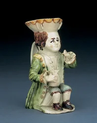 Toby-kande med en drukkenbolt, hans hat med brun og gul tunget kant, hans indridsede brune hår bundet i en pisk og med en hvid roset, siddende med en balusterformet kande på knæet, ca. 1770