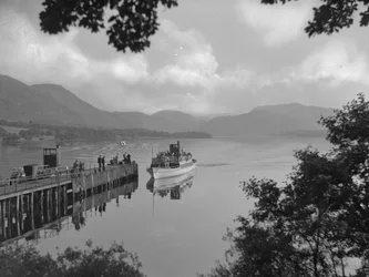 En båd med mennesker på en sø nærmer sig en anløbsbro, fjelde i baggrunden